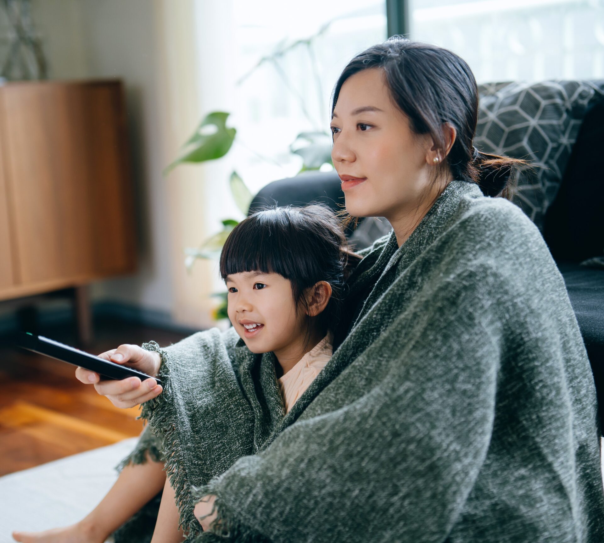Loving young Asian mother and cute little daughter wrapped in blanket, relaxing at home and watching TV in the living room. Bonding between mother and daughter. Family lifestyle. Love and care concept - stock photo