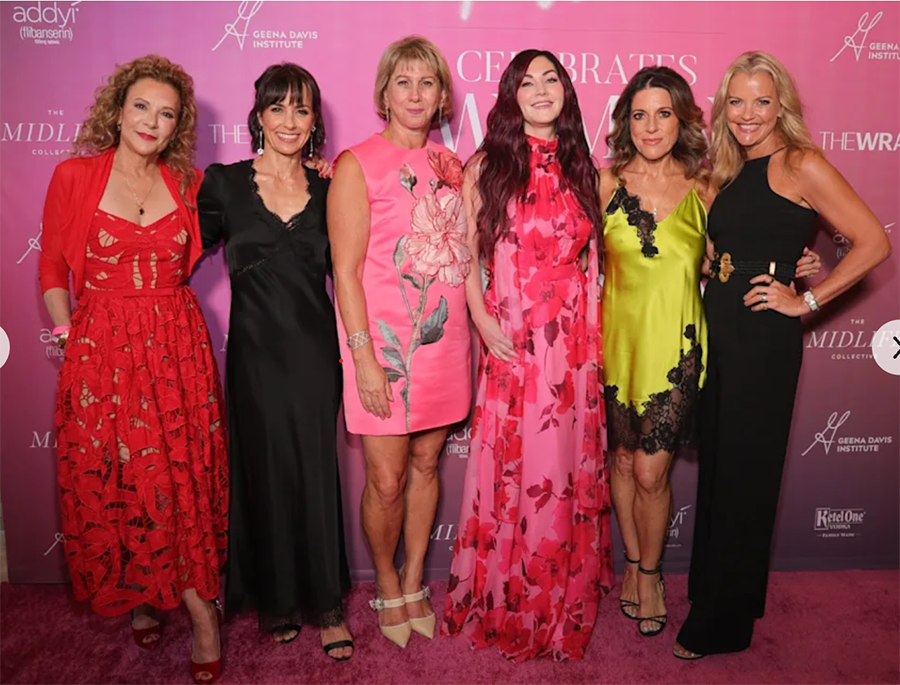 ix women, including Geena Davis Institute CEO Madeline Di Nonno and actress Constance Zimmer, pose together in elegant dresses on a pink carpet in front of a backdrop reading 